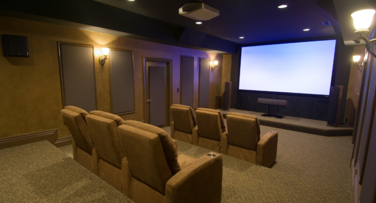 BritePro AV luxury home theater room featuring rows of plush tan recliner seats, a large projection screen, ceiling-mounted projector, surround sound speakers, and elegant wall sconce lighting in a professionally designed private cinema setup.