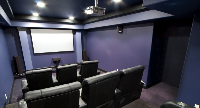 BritePro AV custom home theater featuring rows of black leather recliner seats with built-in cup holders, a ceiling-mounted projector such as Sony or JVC, a Stewart or Elite Screens projection screen, floor-standing speakers compatible with JBL Synthesis or Revel surround sound systems, and recessed lighting set against deep blue walls for a fully immersive private cinema experience.
