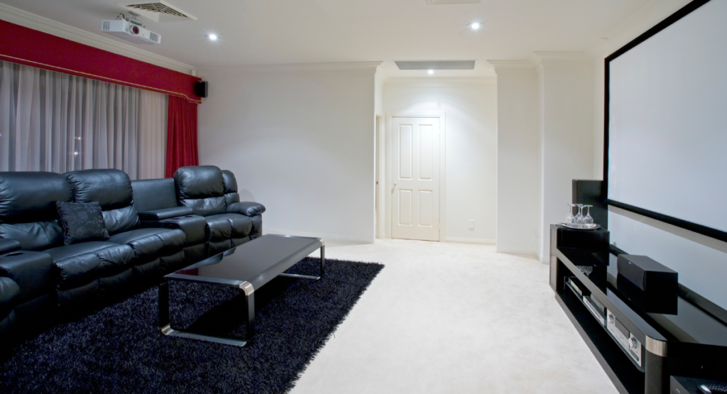 Image 1 Alt Text: "BritePro AV elegant home theater room featuring a black leather recliner sofa, sleek coffee table on a dark shag rug, ceiling-mounted projector such as Epson or BenQ, a large Stewart or Screen Innovations projection screen, and a media console housing Marantz or Denon AV receivers, set in a bright, clean room with red drape accents and recessed lighting.